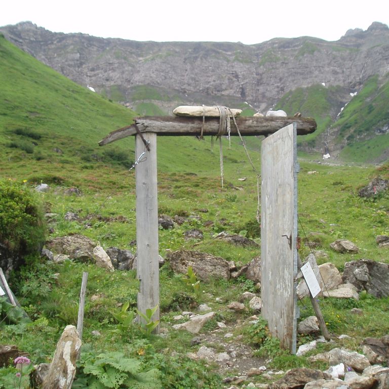 Inmitten einer Gebirgslandschaft steht ein Türrahmen mit geöffneter Tür. Ein schmaler Pfad schlängelt sich hindurch